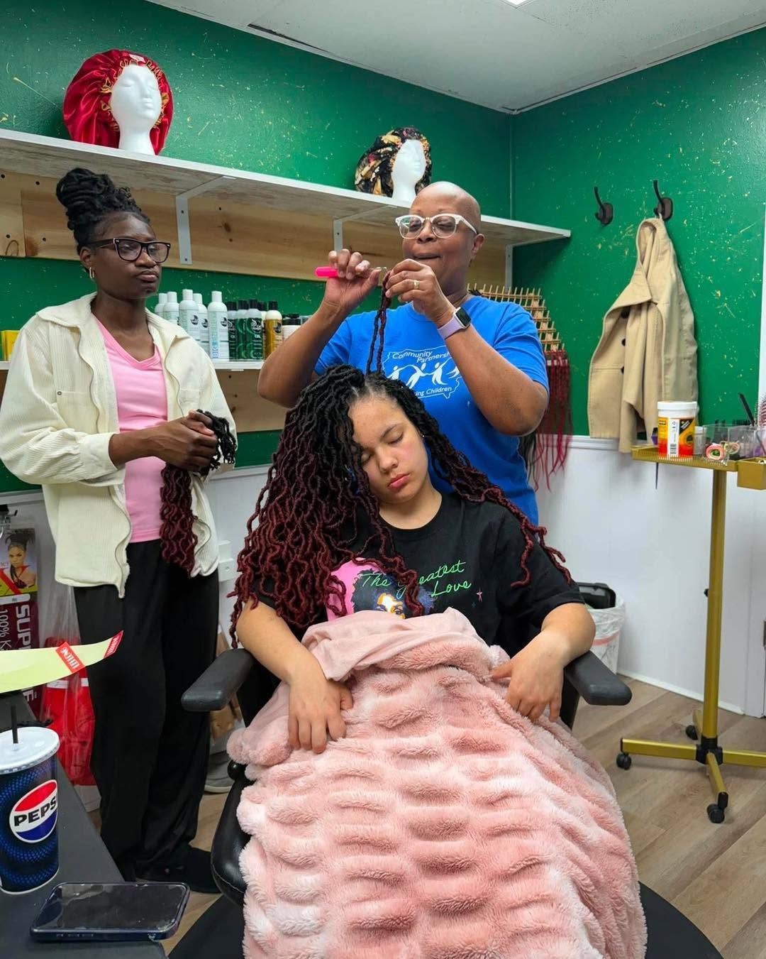 Salon scene with stylist braiding a seated client’s hair while another person watches nearby.