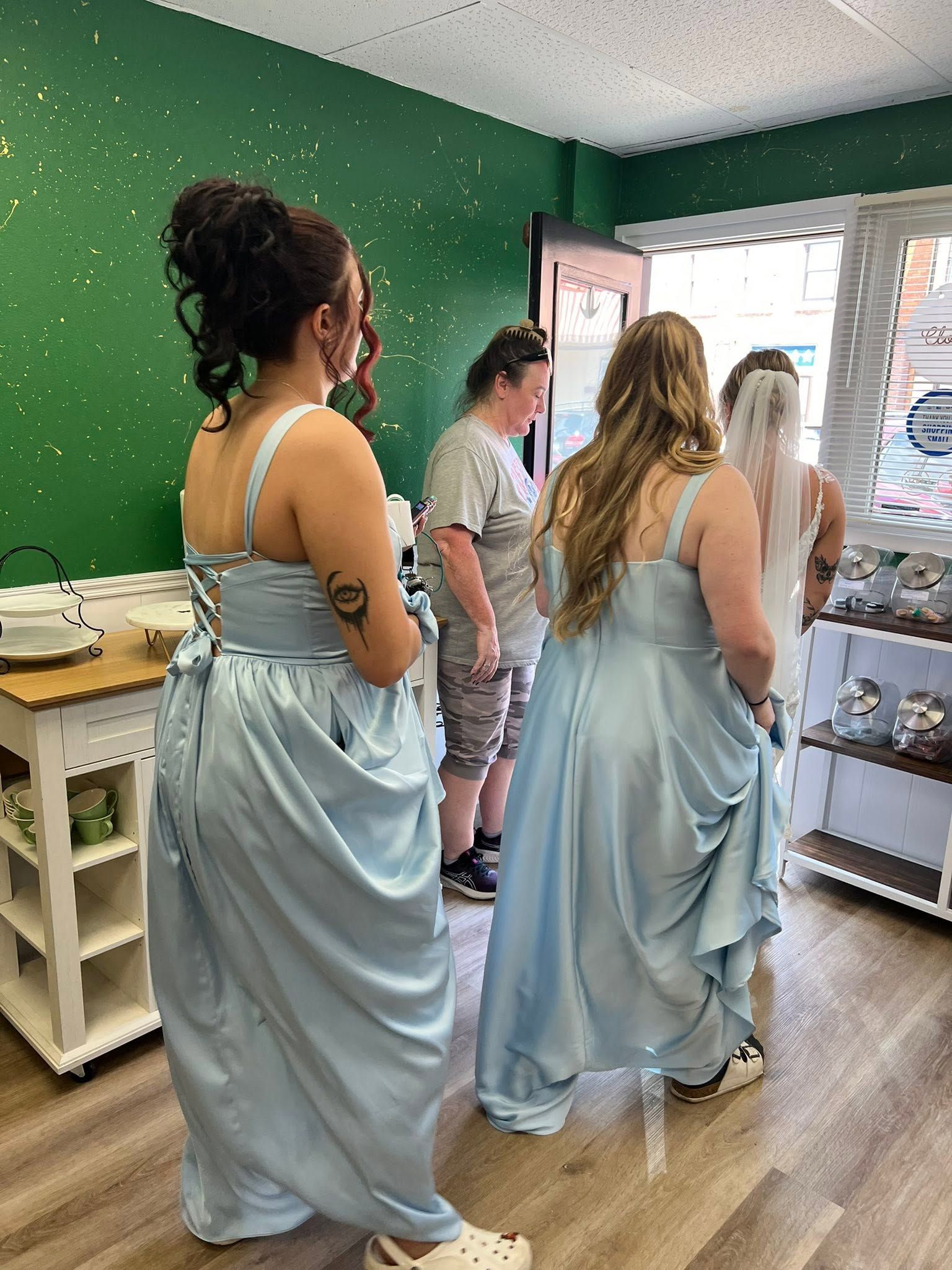 People in light blue gowns standing in a room with green walls, near a window and folded fabrics.