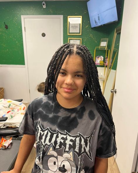 Smiling person with braided hair wearing a black graphic T-shirt in a room with green walls and a wall-mounted TV.