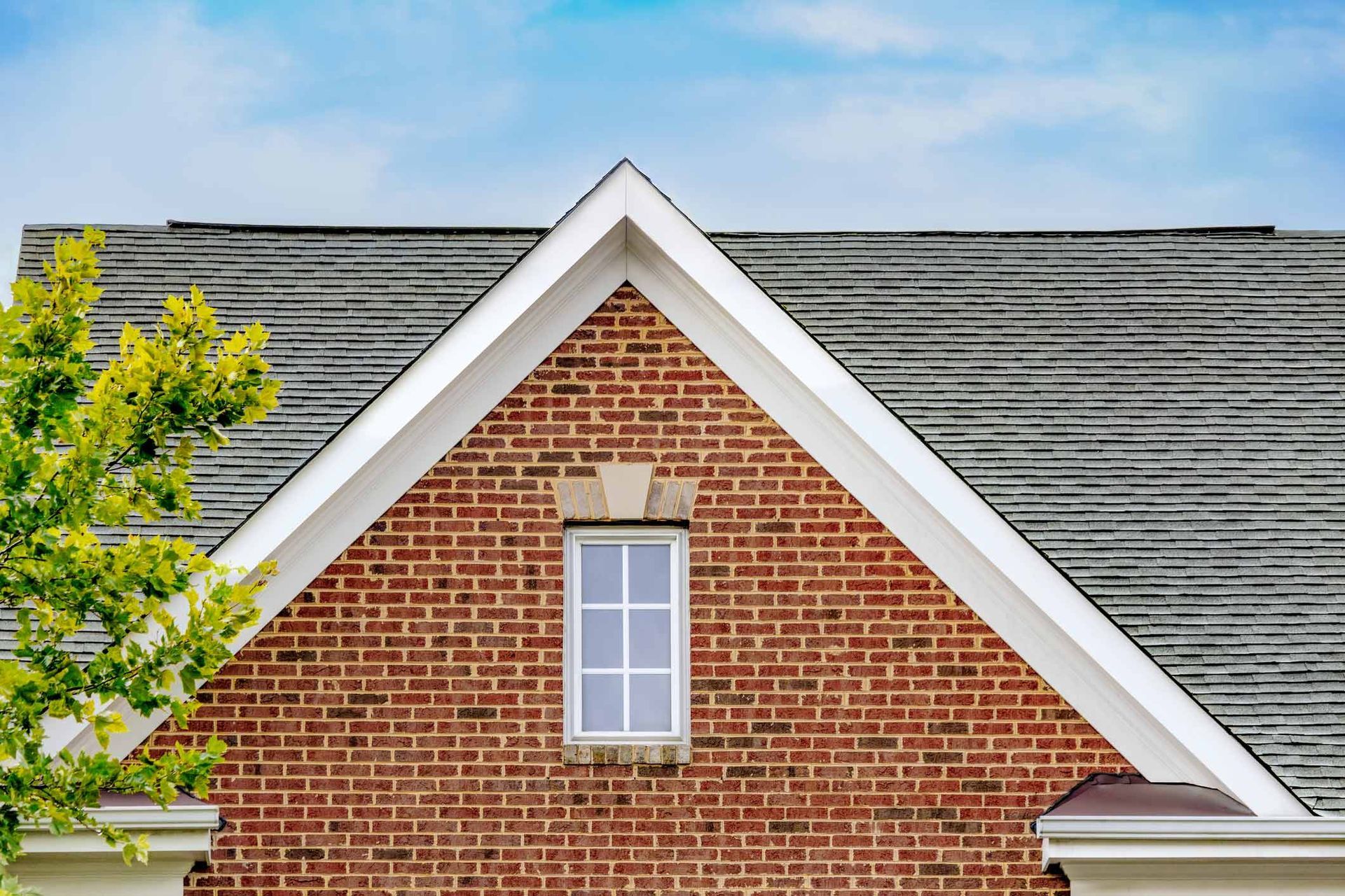 Brick gable with white-trimmed window, gray roof, and tree against a blue sky.