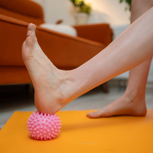 Foot pressing a pink massage ball on an orange yoga mat in a living room, suggesting heel pain treatment