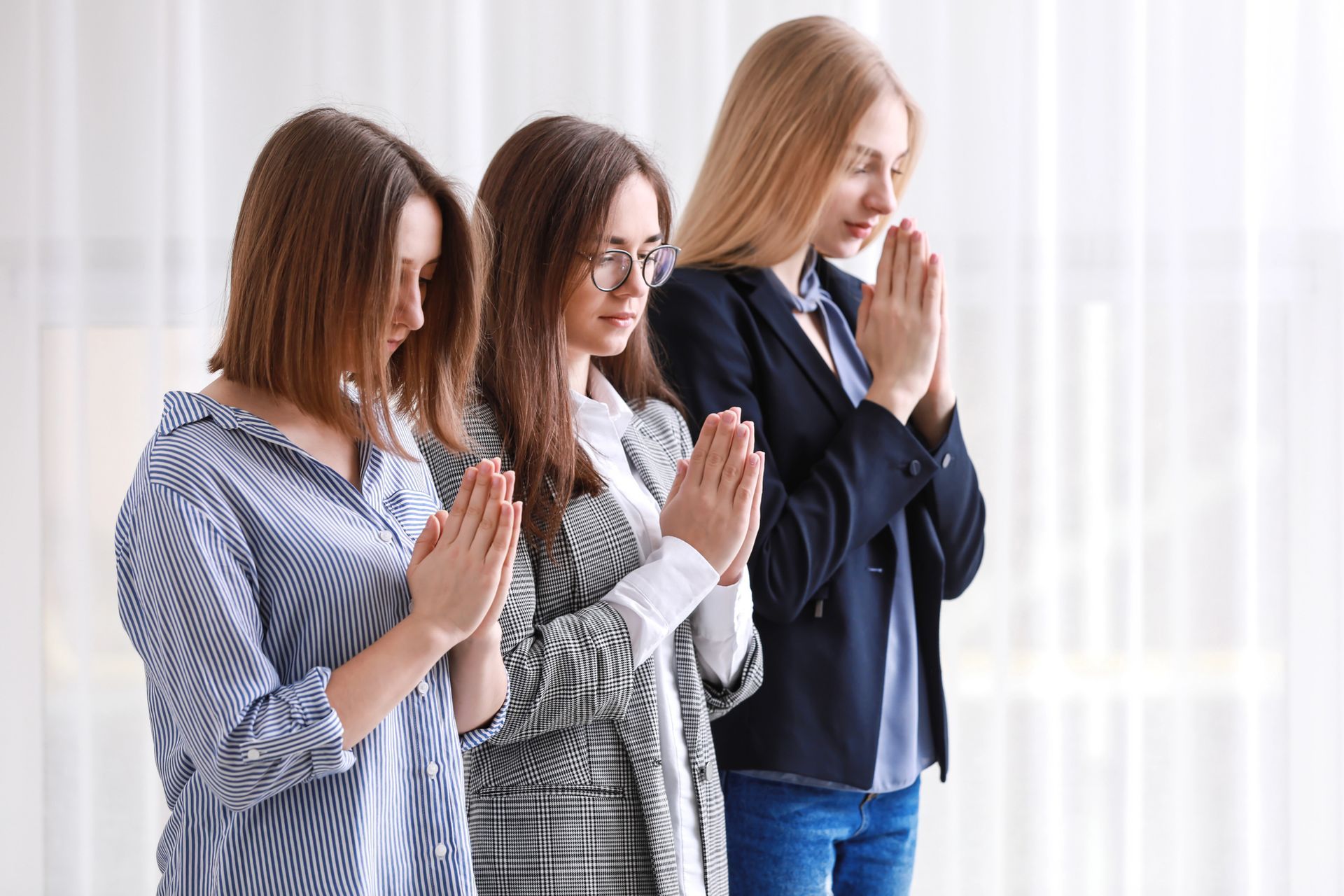 Tres mujeres permanecen de pie con las manos juntas en oración y los ojos cerrados en una habitación bien iluminada.