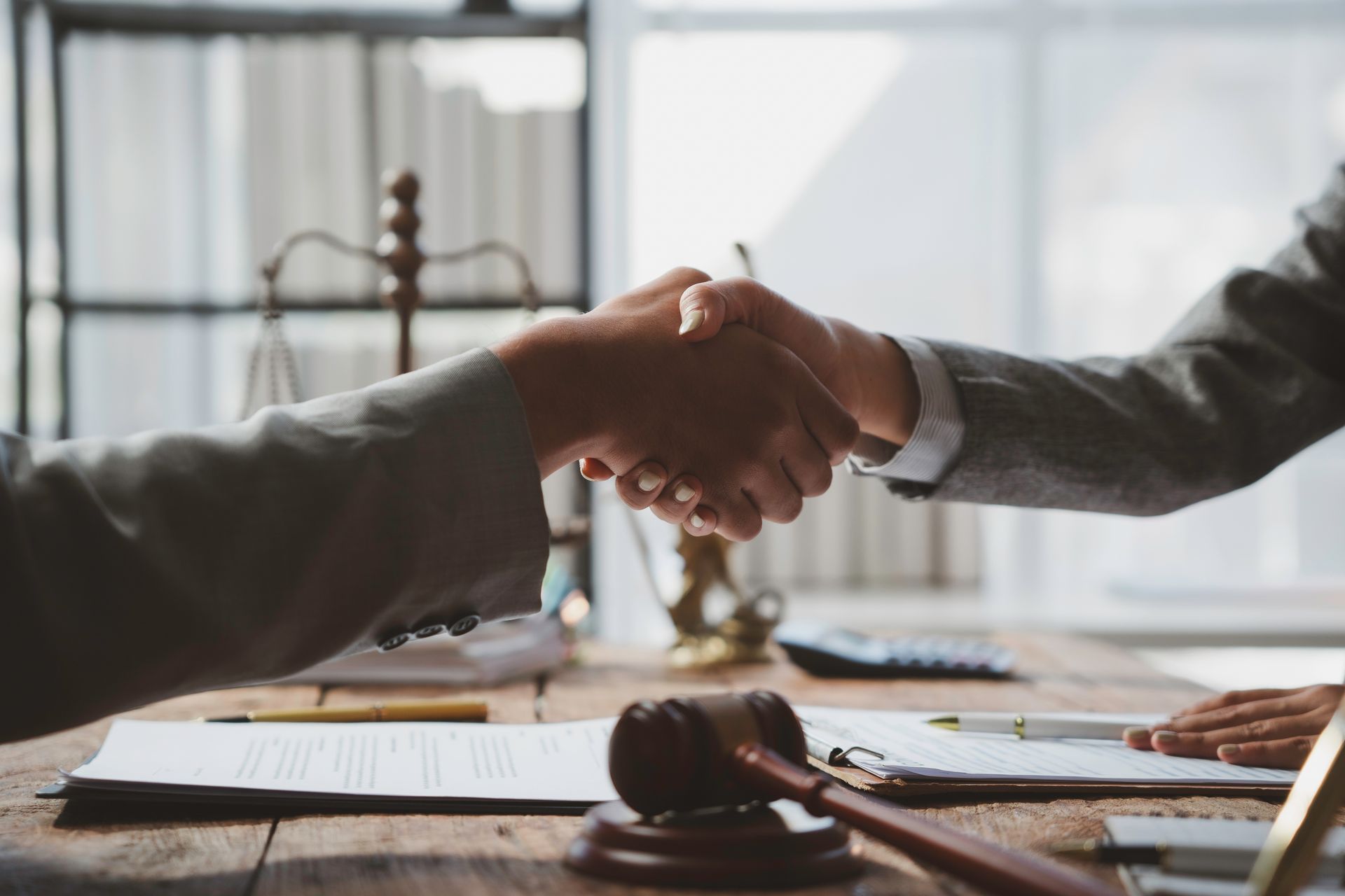 Two people in business attire shake hands across a desk with documents and a gavel, suggesting a legal agreement.