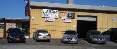 Four parked cars outside a yellow auto repair shop with signs on the front wall. | A-Tech Auto Repair