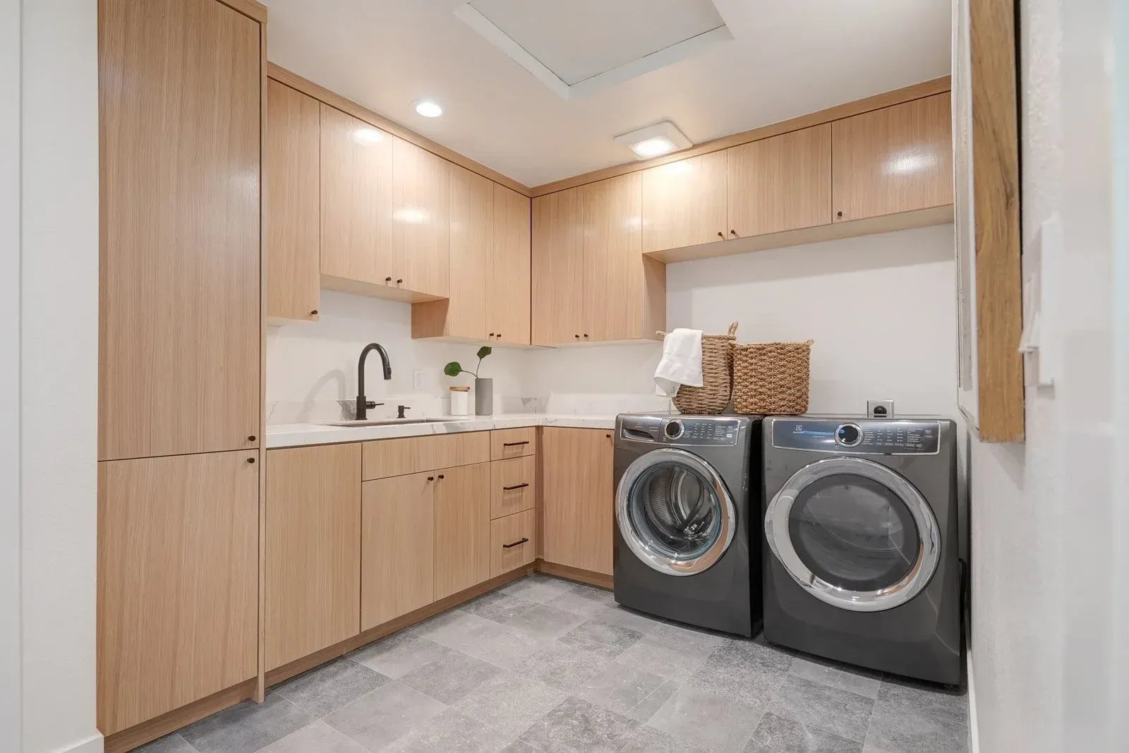 Laundry room with light wood cabinets, dark appliances, white countertops, and gray floor tiles.