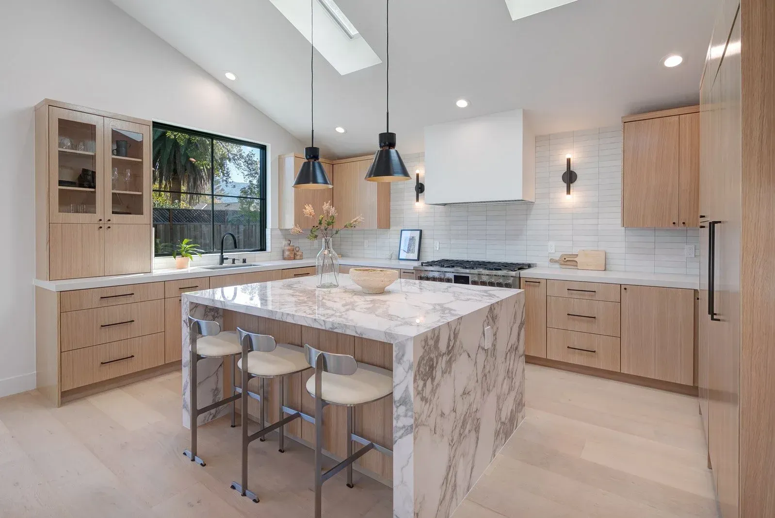 Modern kitchen with light wood cabinets, island with marble countertop, and three stools.