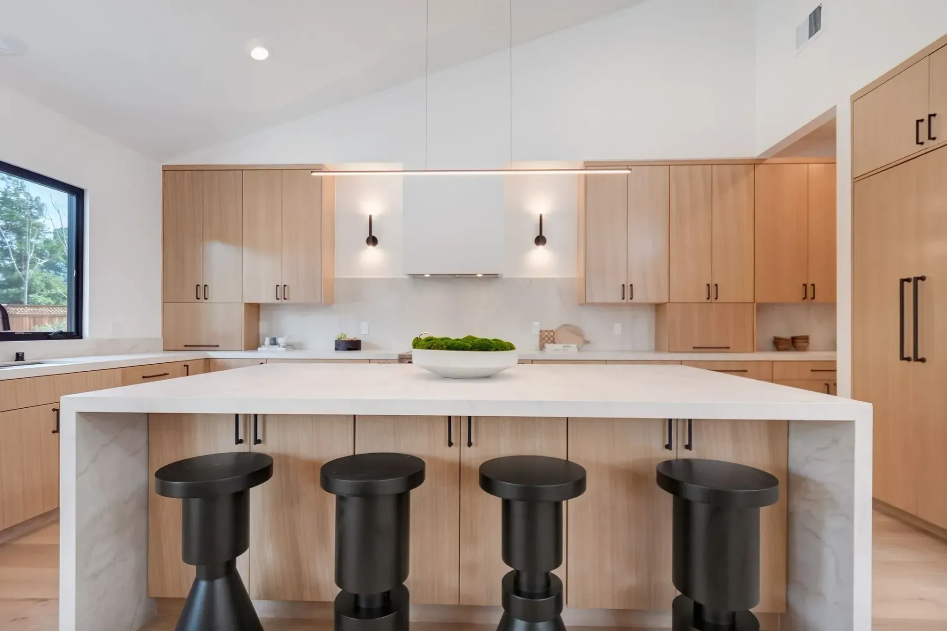 Modern kitchen with light wood cabinets, white countertops, and black stools at an island.