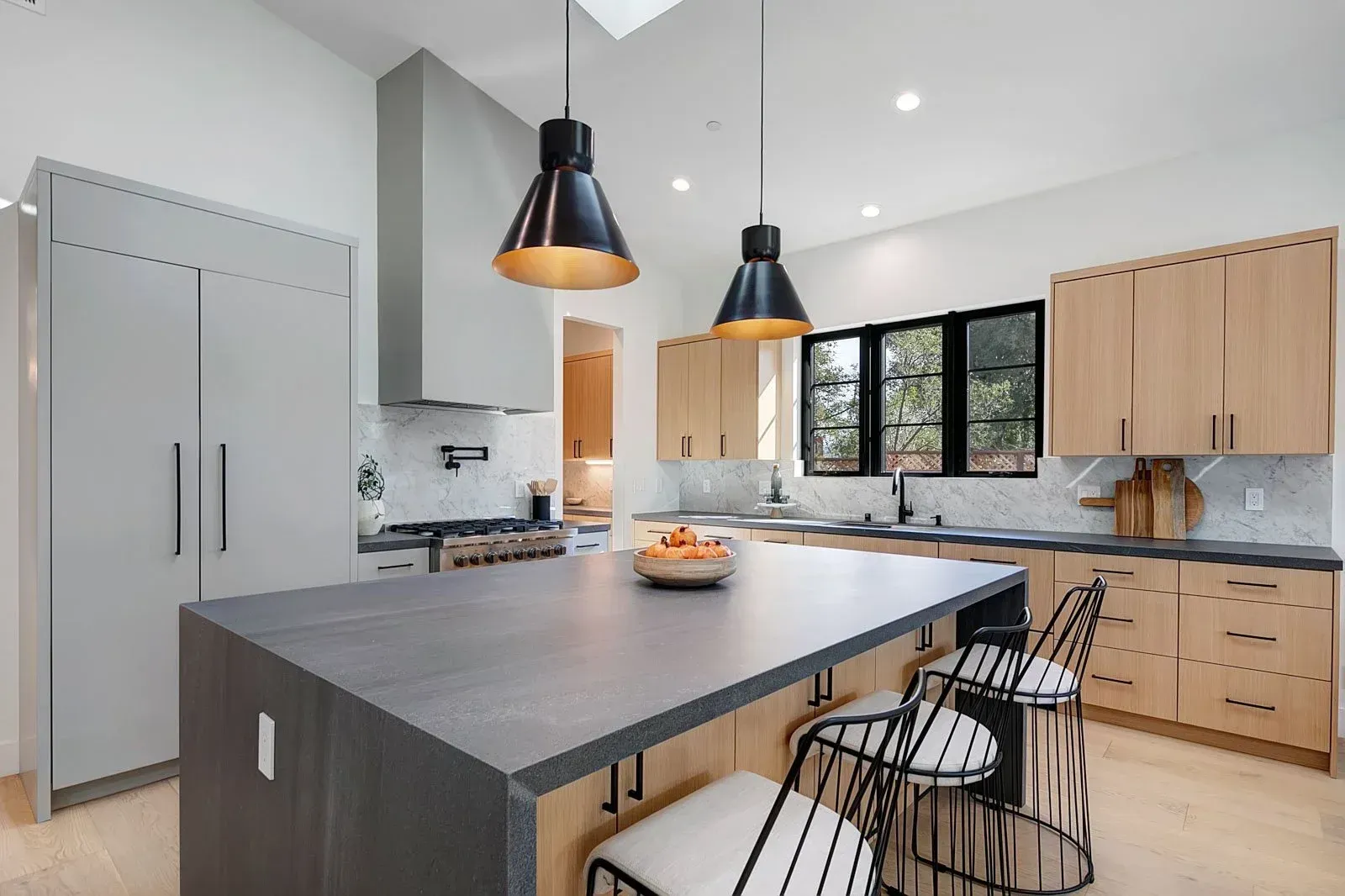 Modern kitchen with island, pendant lights, stainless steel hood, light wood cabinets.