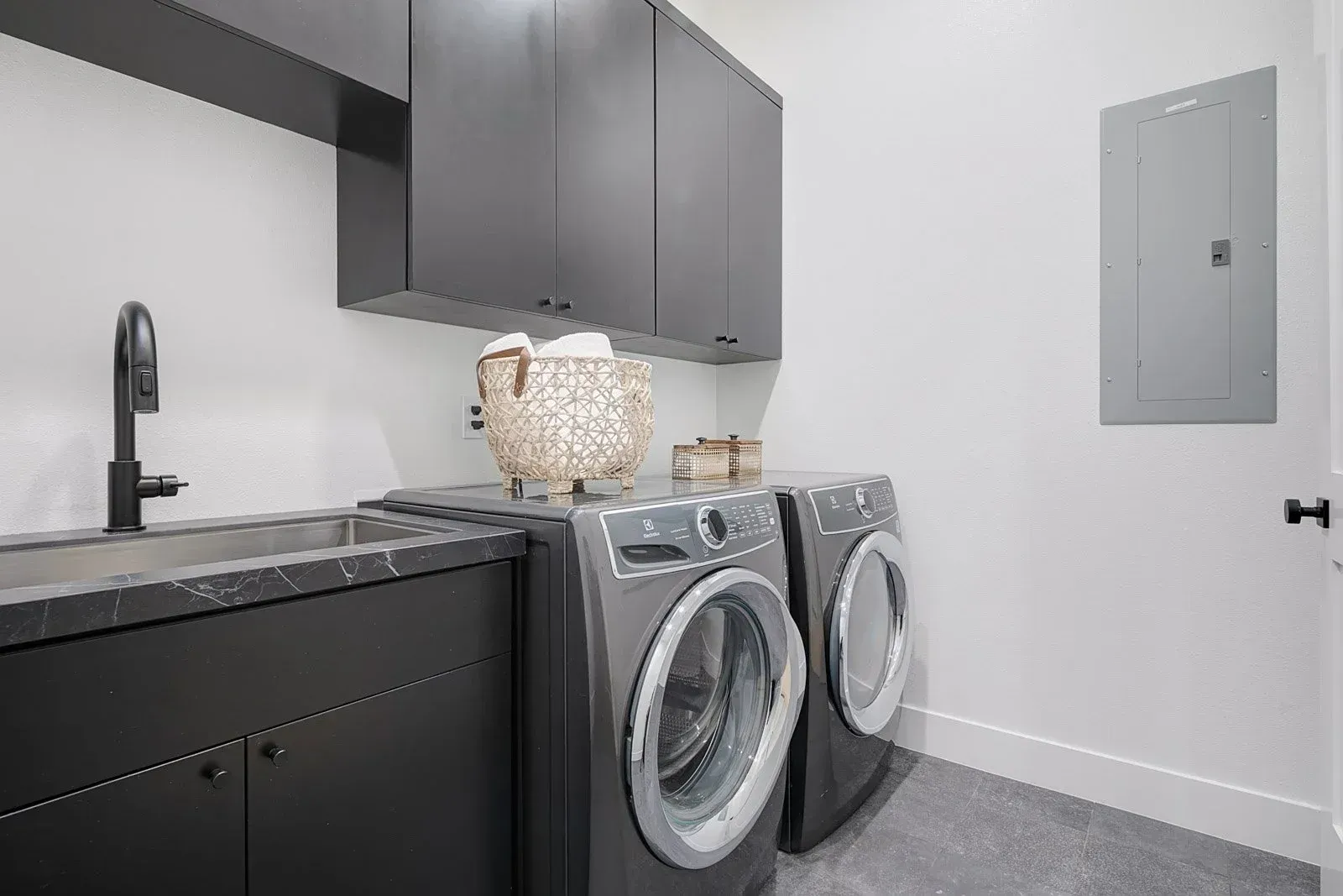 Modern laundry room with dark cabinets, gray washer/dryer, sink, and electrical panel.