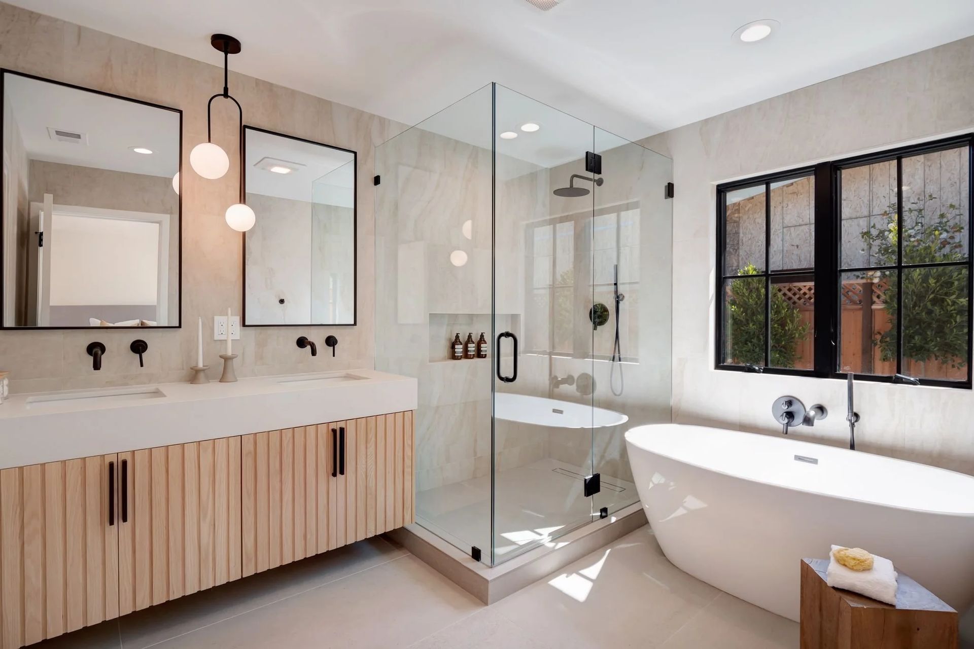 Modern bathroom: double vanity with wood cabinets, glass shower, soaking tub, black-framed mirrors, and window.