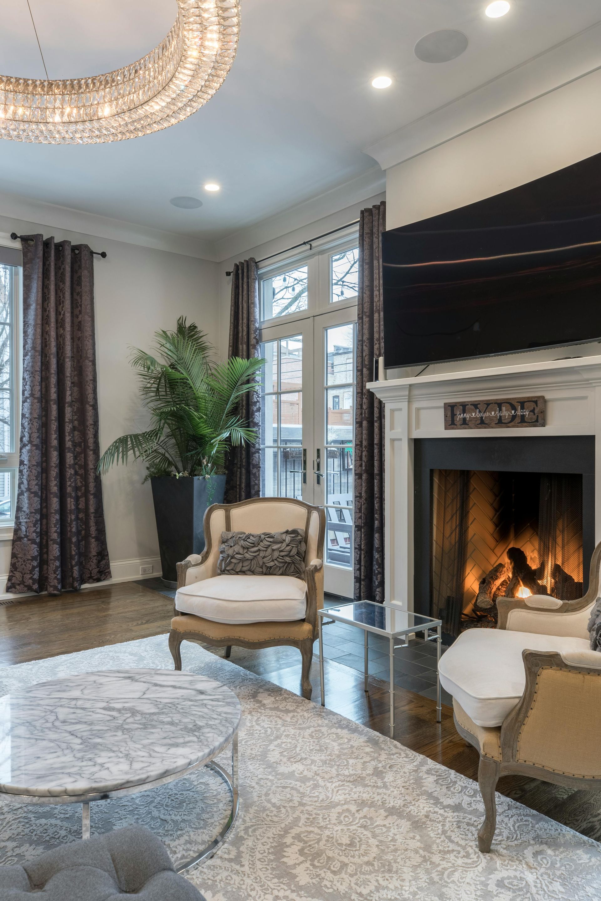 Living room with fireplace, chairs, round rug, large chandelier, and French doors.