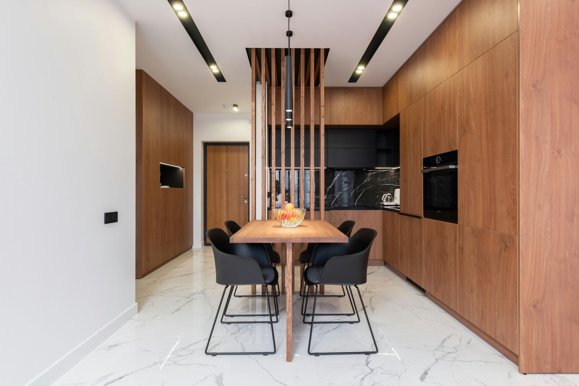 Modern kitchen with wood cabinetry, marble floor, dining table, and black chairs.