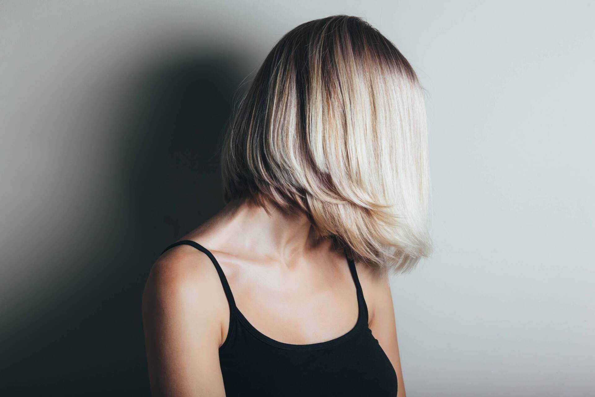 Persona con capelli biondi di media lunghezza, con indosso una canottiera nera, di spalle, su sfondo grigio.
