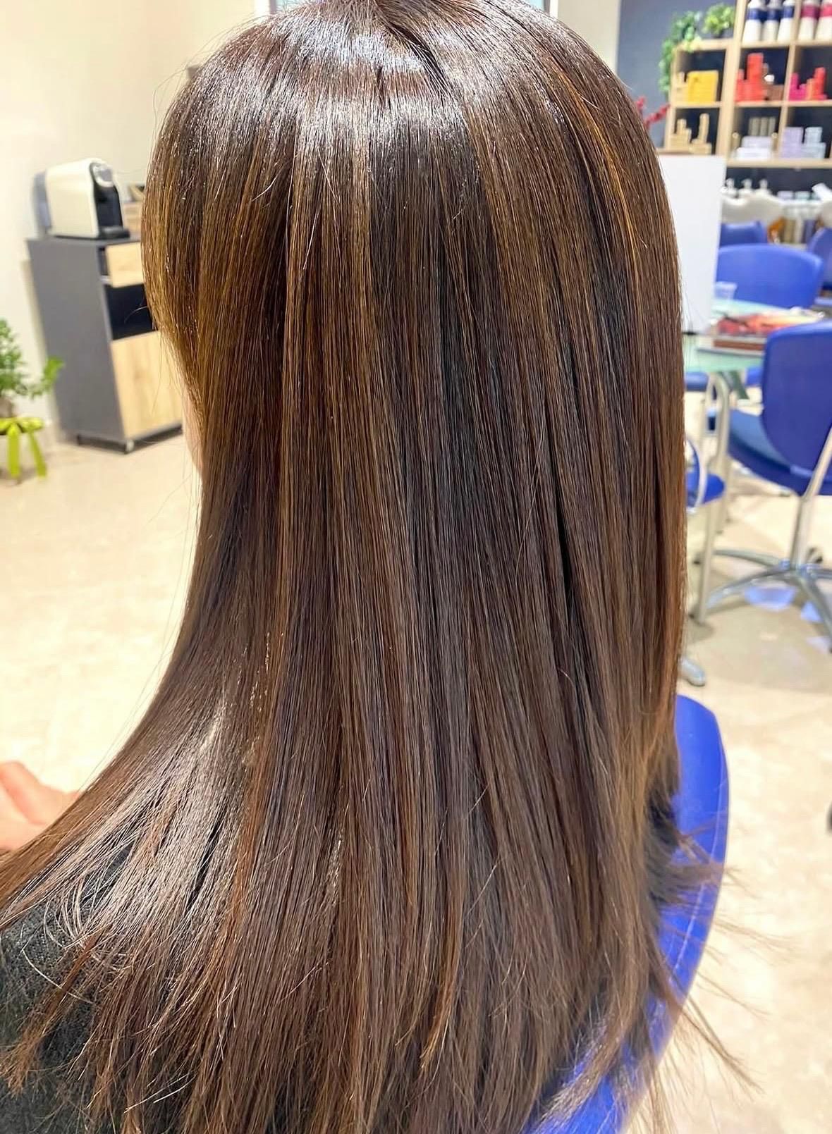Lunghi capelli castani lisci con sottili riflessi, seduti su una poltrona da parrucchiere, visti da dietro.