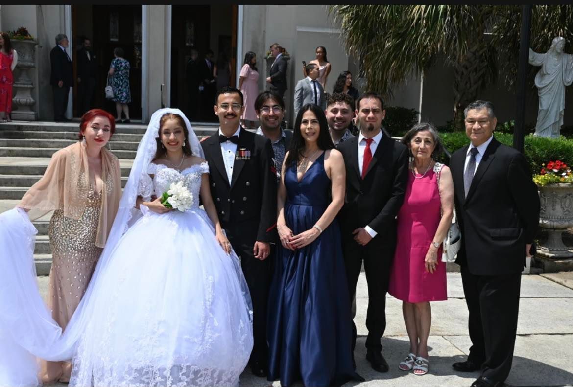 Wedding group outside a church; bride in white dress, veil, and bouquet; groom in navy suit; guests in formal attire.
