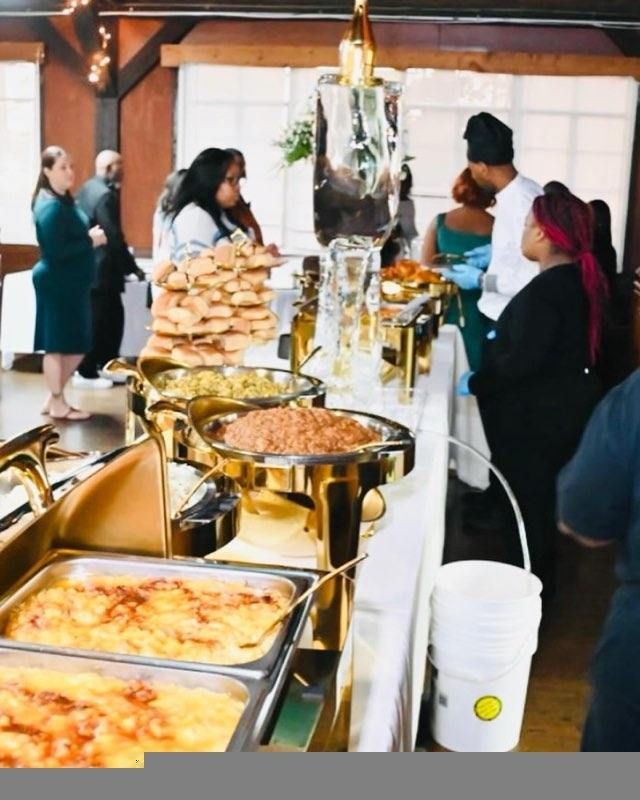 A buffet table with various dishes, attended by guests and a chef in a black hat. The setting appears to be a social event.