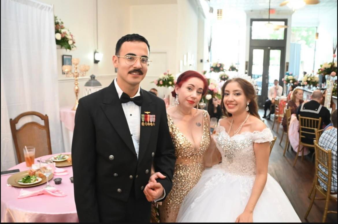 A man in a naval uniform poses with two women in formal dresses at an event. The setting appears to be indoors, likely a banquet hall.