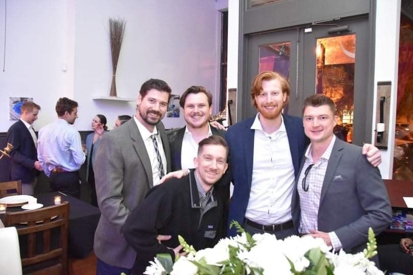Five men smile and pose for a photo in a restaurant. Two men in the background are also visible.