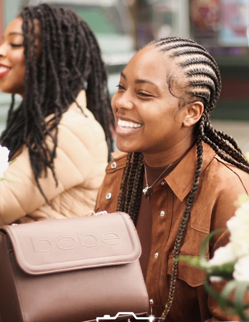 Woman with braided hair smiles, wearing a brown jacket, sitting with a friend at a cafe. A brown handbag sits on the table.