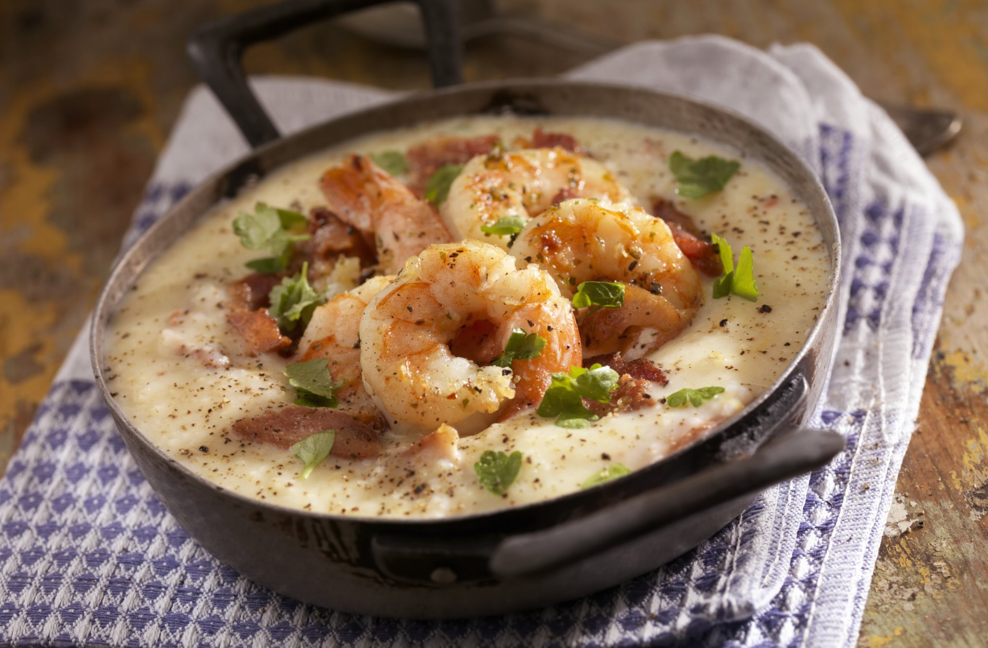 Shrimp and grits in a dark bowl, garnished with parsley and bacon, on a checkered cloth.