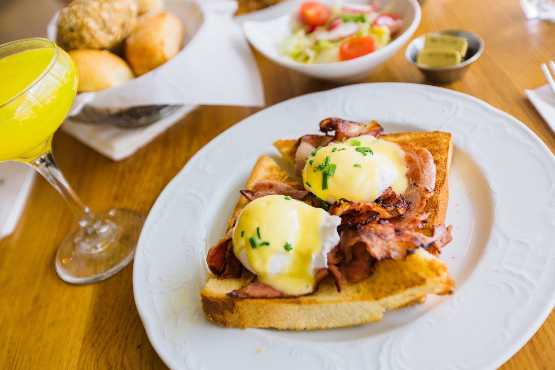 Eggs Benedict on toast with bacon, a side salad, bread rolls, and a glass of orange juice on a wooden table.