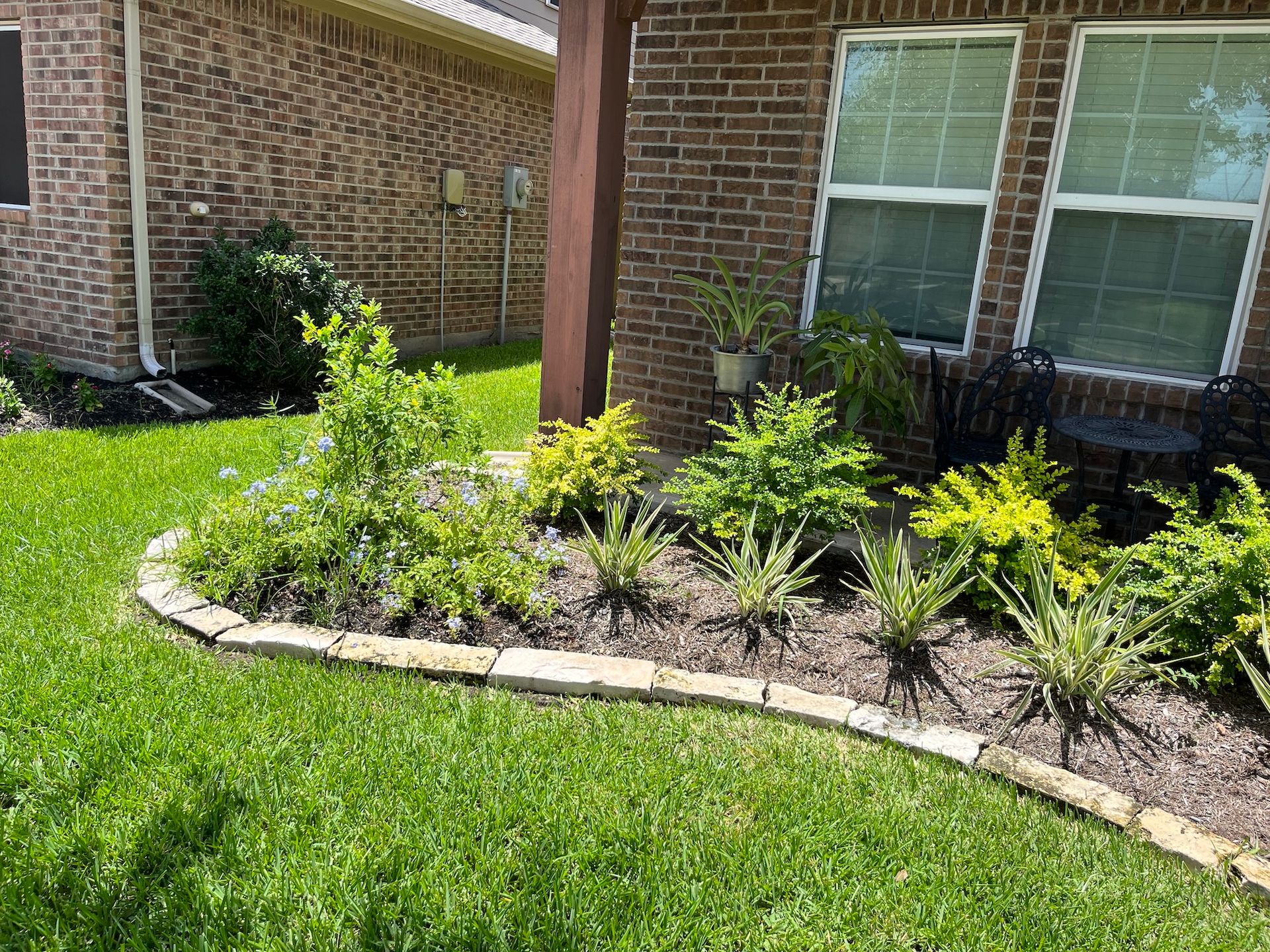 Landscaped flower bed with various green plants and stone edging in front of a brick building.