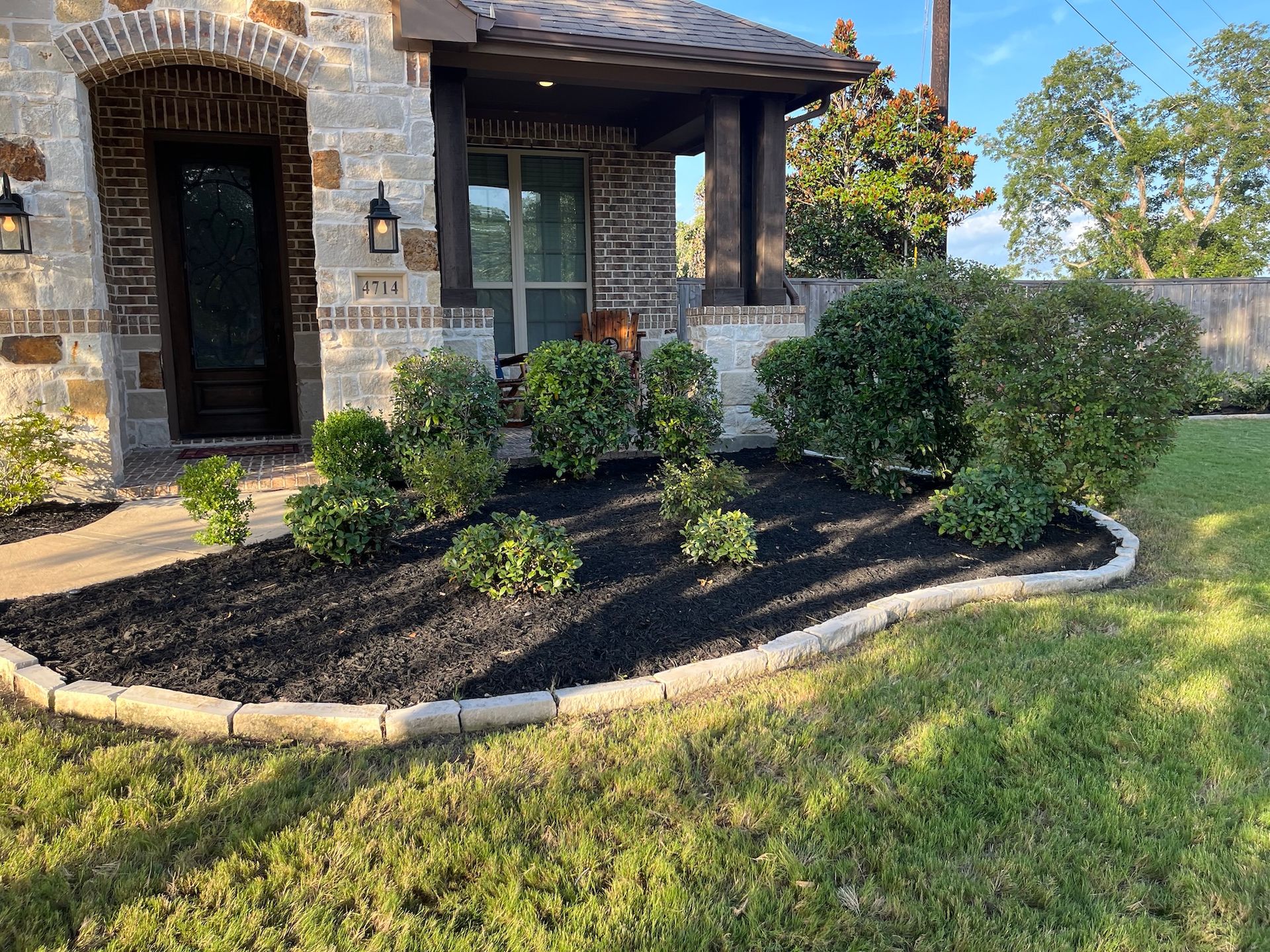 Well-manicured front yard with dark mulch, green bushes, and stone house.