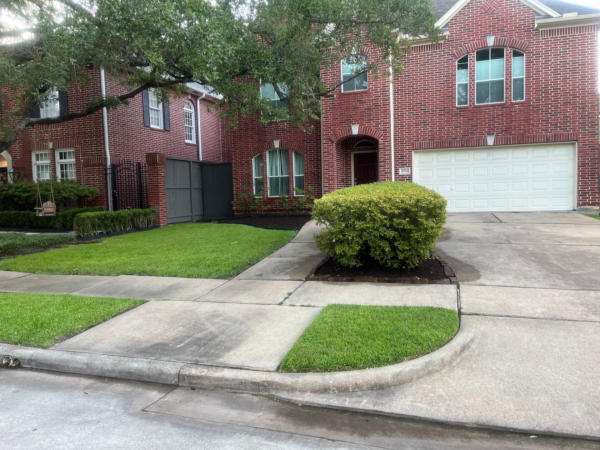 Brick house with garage, lawn, and sidewalk. Green bushes and trees in front.