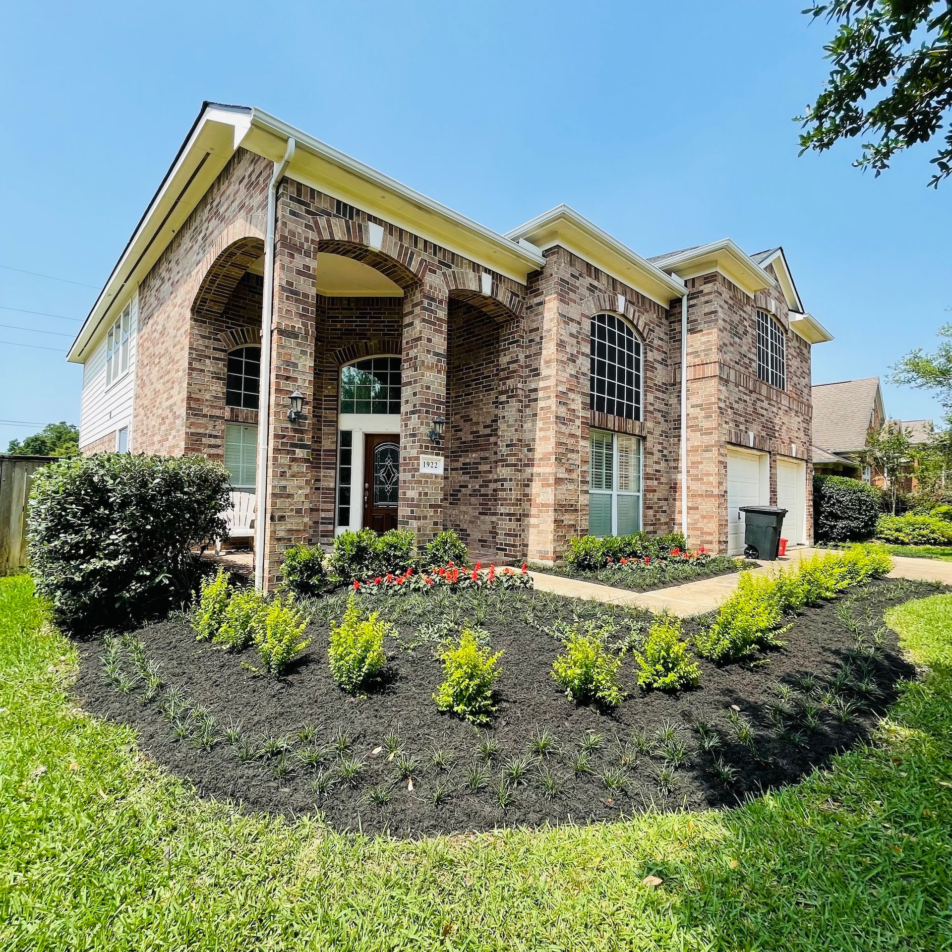 Two-story brick house with arched entrance, landscaped yard, and blue sky.