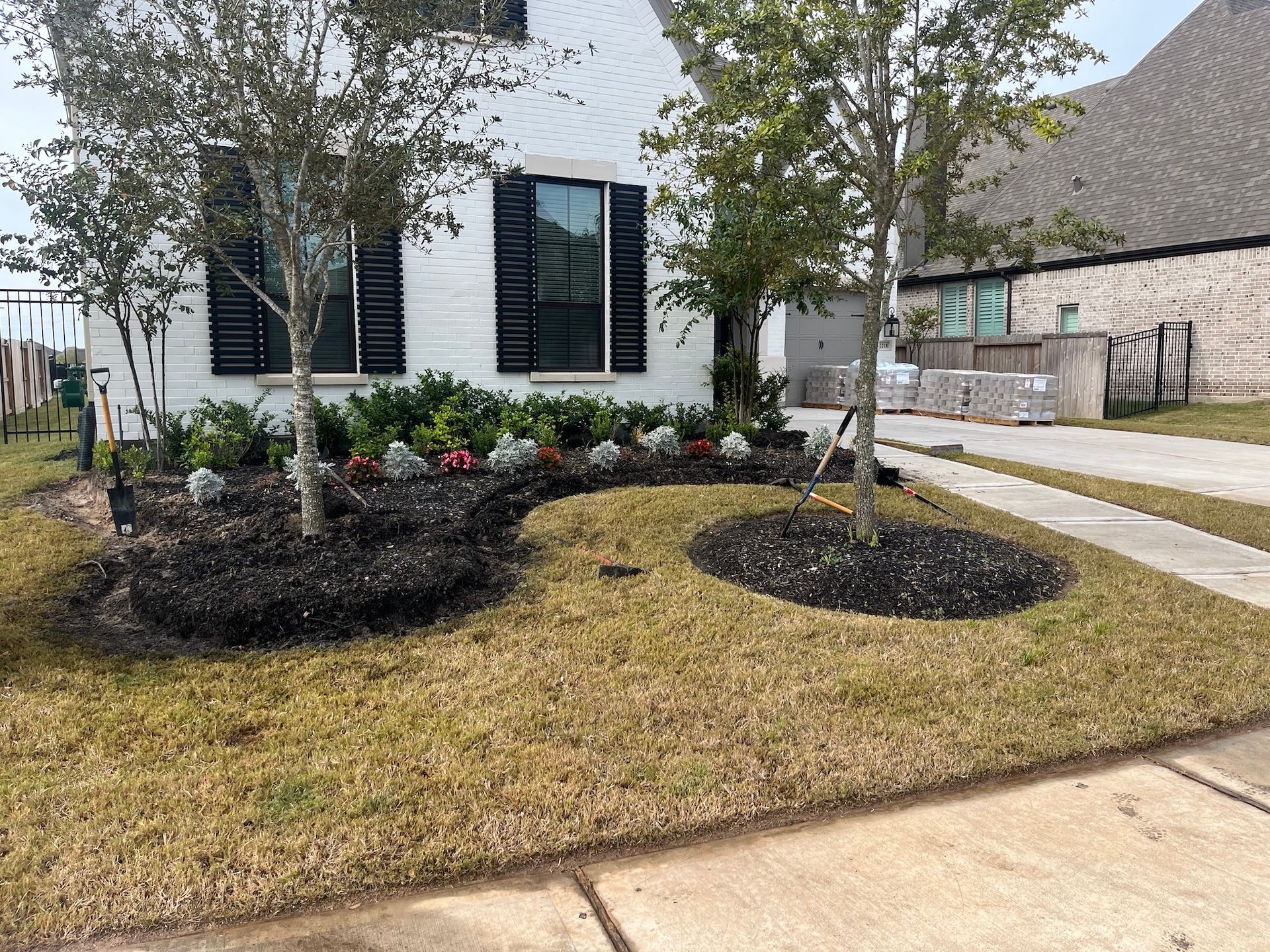 White house with a landscaped yard; trees, dark mulch, green and red flowers, and brown grass.