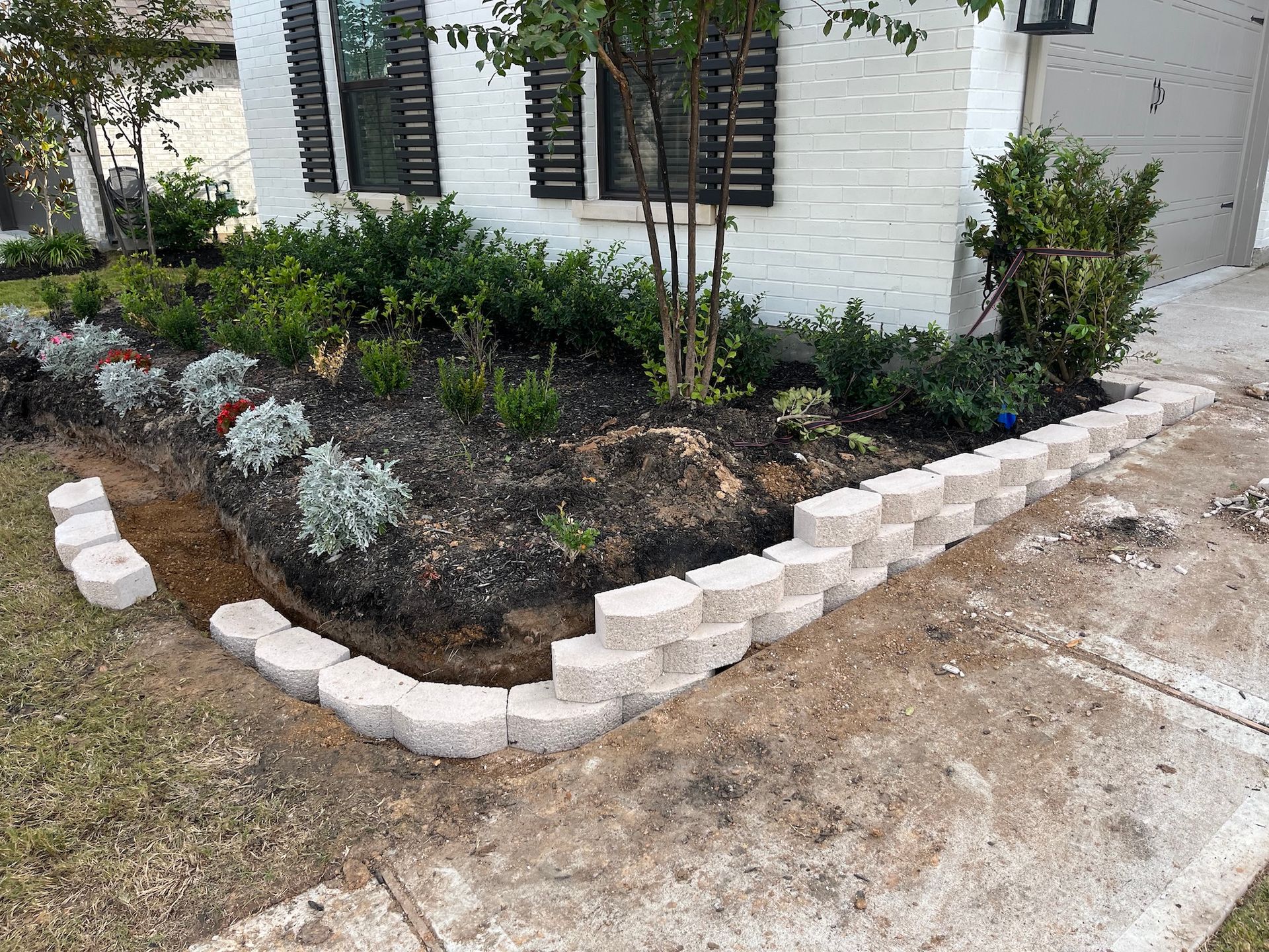 Landscaped garden bed with retaining wall. Flowers and shrubs in front of a white brick house.