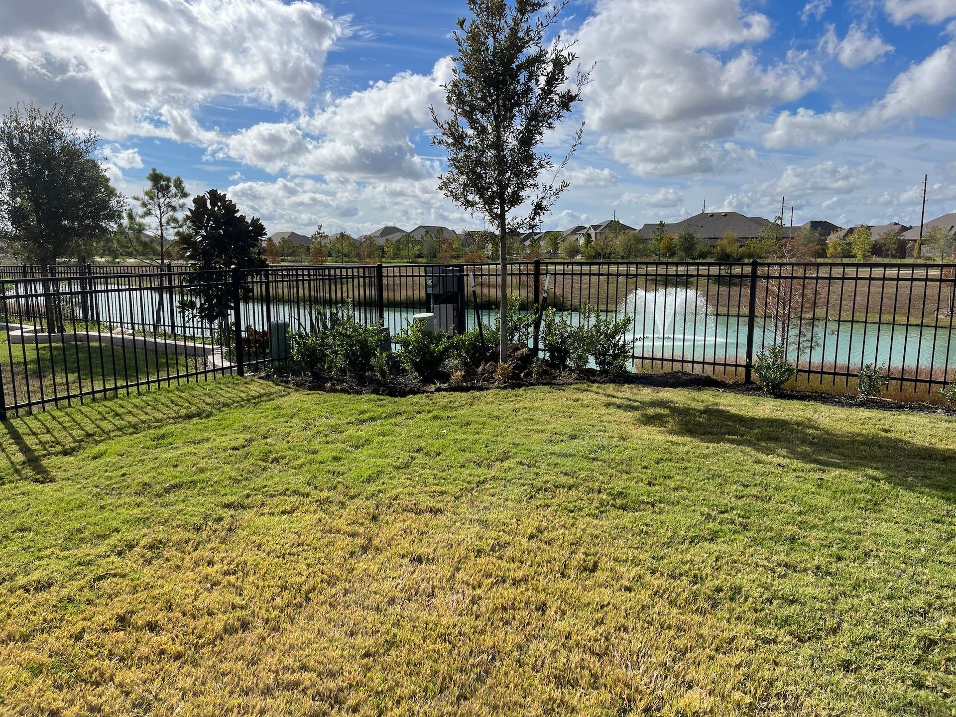 Grassy yard with a dark fence overlooking a pond under a cloudy blue sky.