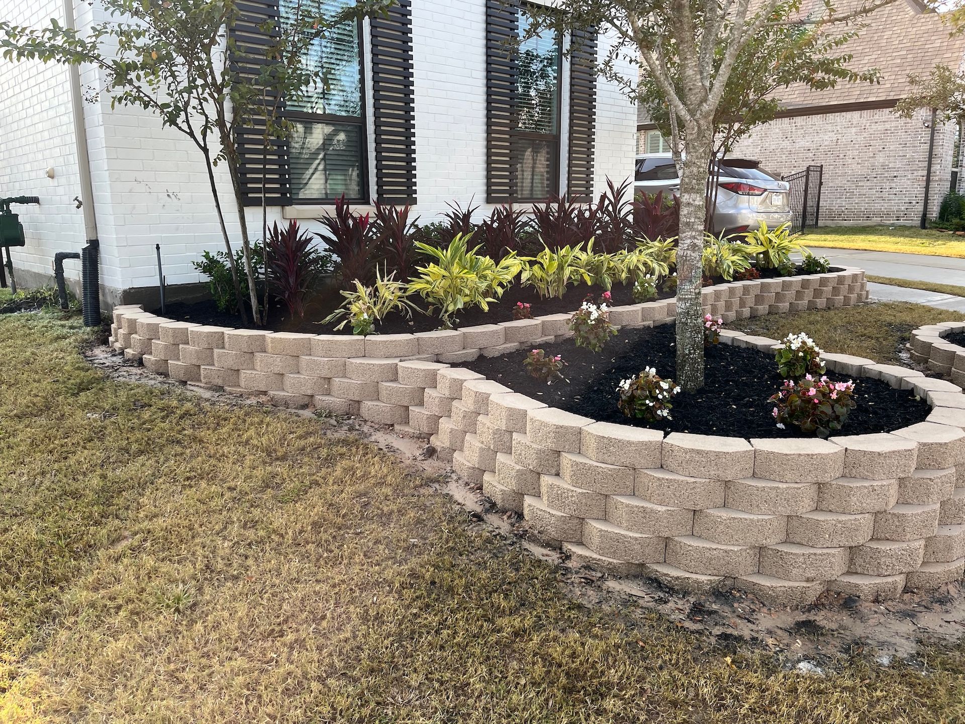 Landscaped yard with retaining wall, flower beds, and a tree in front of a house.