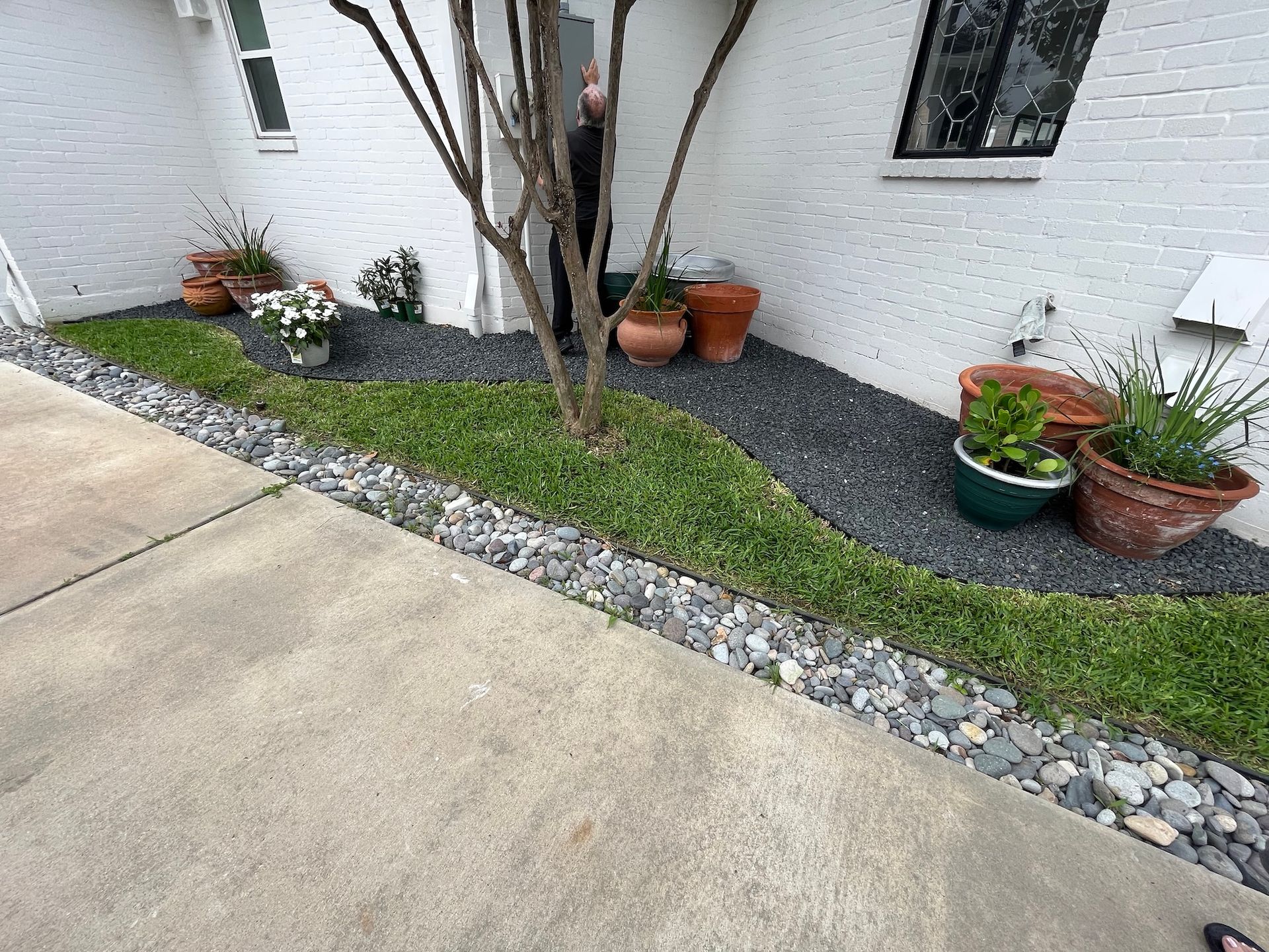 Landscaped area with grass, gray rocks, and potted plants against a white brick building.