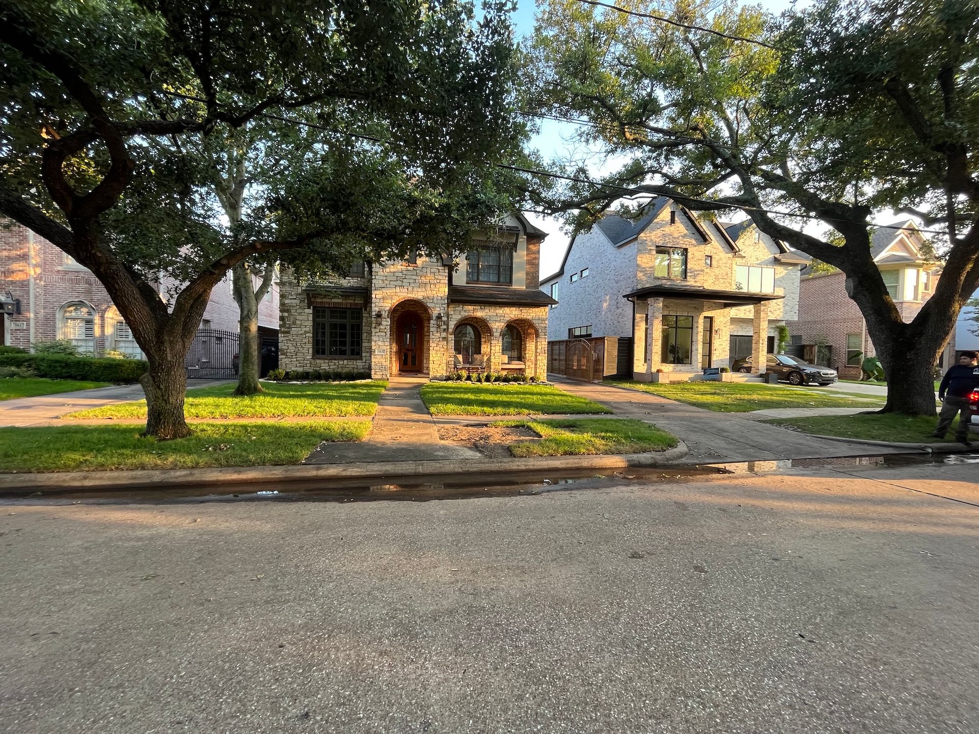 Stone houses with large trees in front, viewed from the street. One person stands on the right.