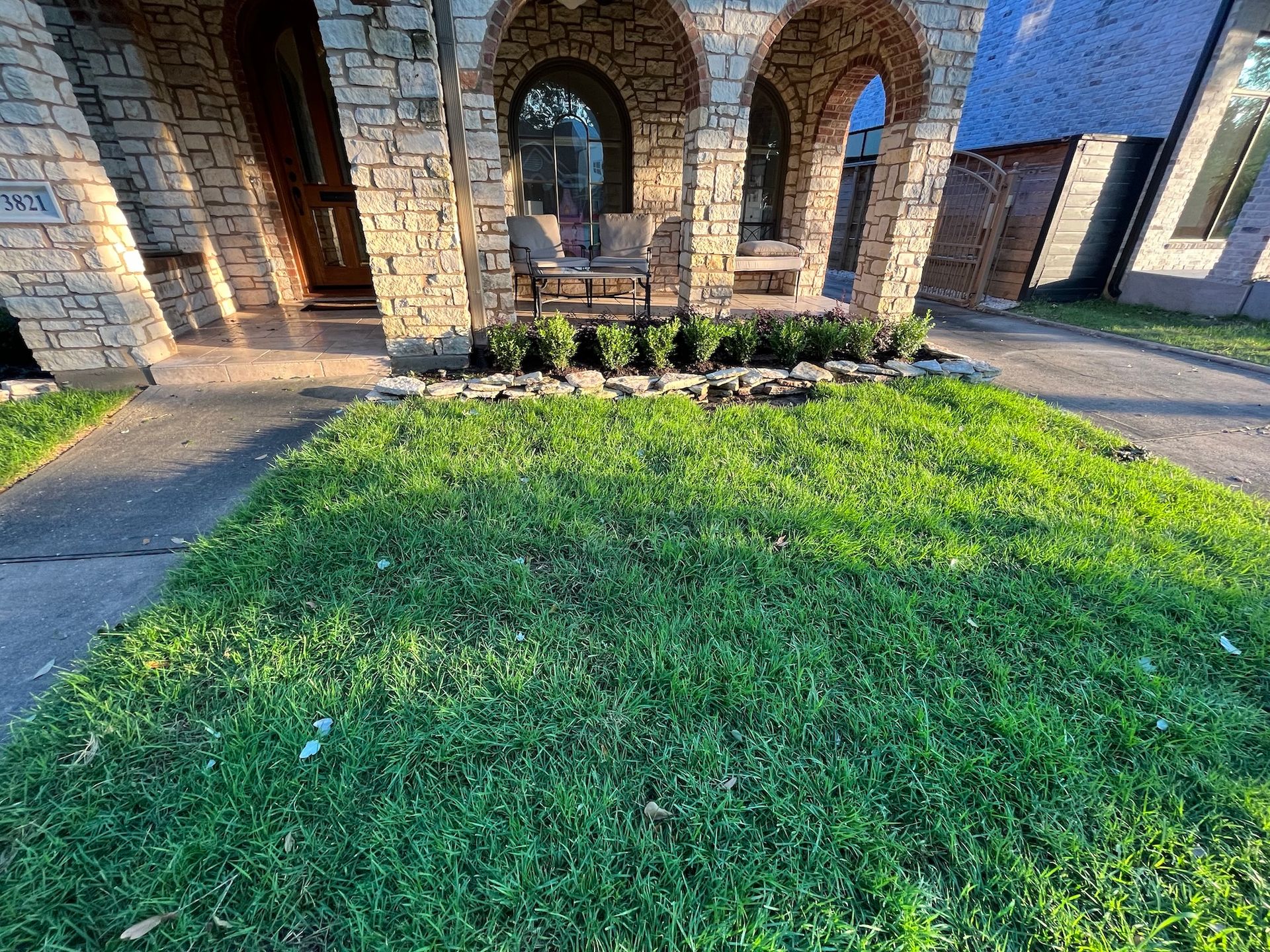 Green lawn in front of a house with stone arches, a porch with seating, and a small hedge.