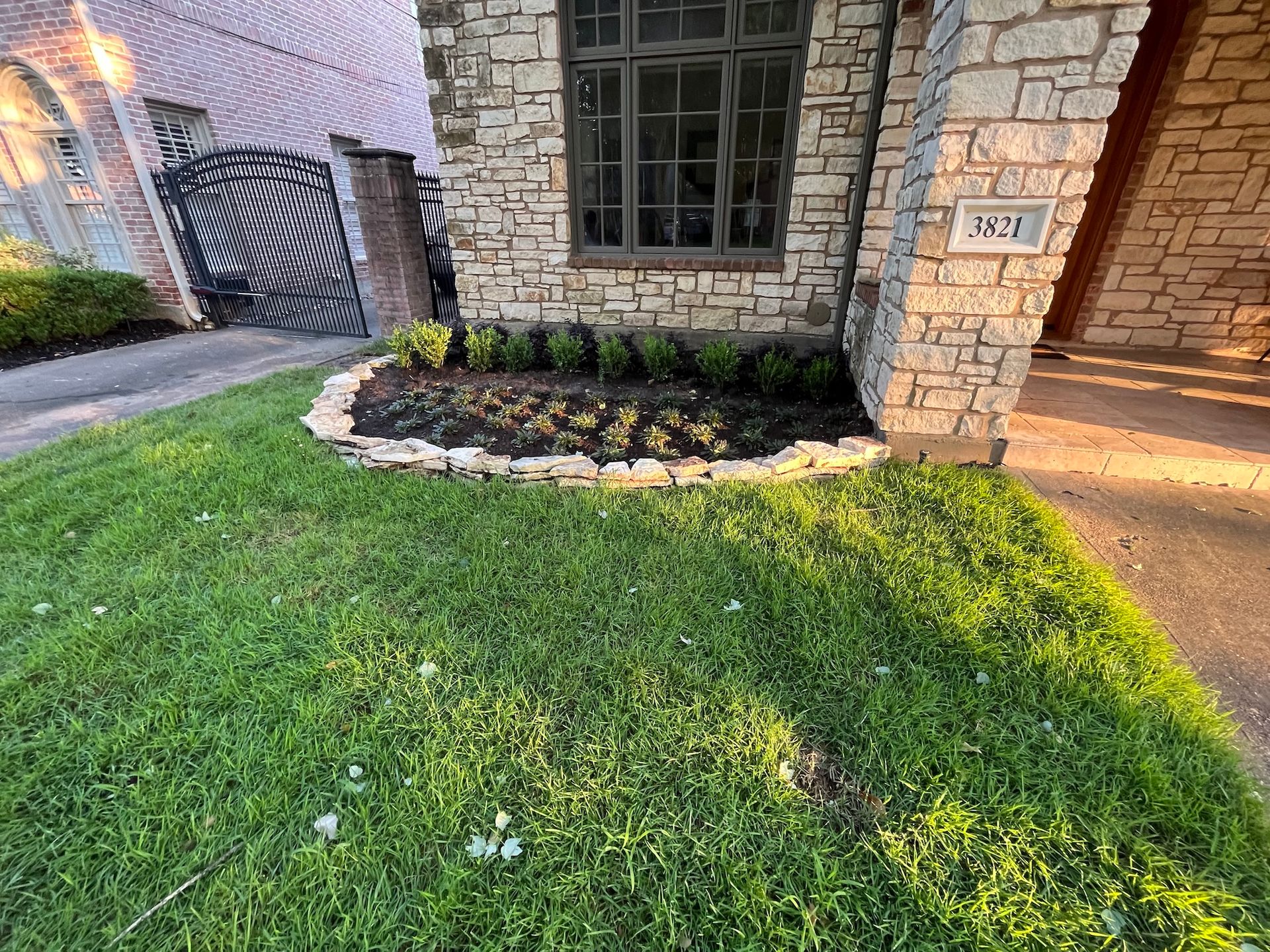 Lawn and flowerbed in front of a stone house with a dark window. The yard has green grass and a paved driveway.