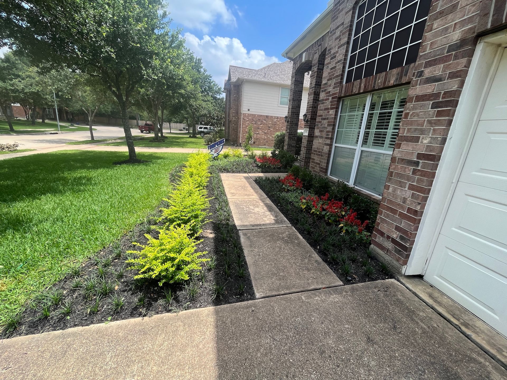 A brick house with a walkway, surrounded by greenery and flowers under a blue sky.