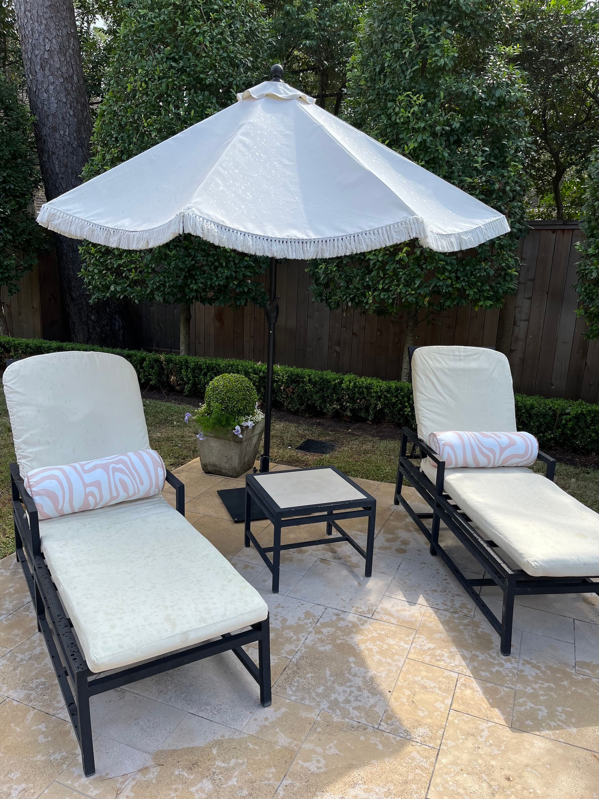 Two cream-cushioned lounge chairs under a white umbrella on a patio with a small table, surrounded by greenery.