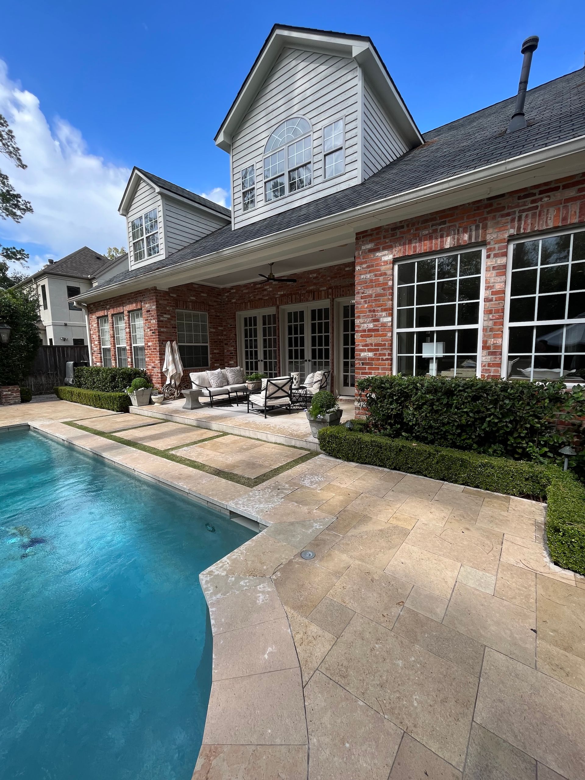 Backyard with pool, patio, and brick house. Blue water, beige stone, green hedges, white furniture, and a sunny sky.