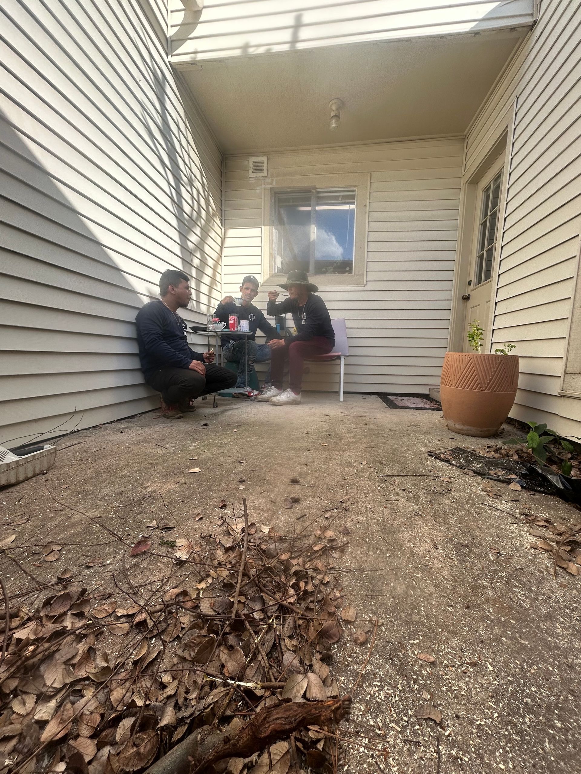 Three people seated on a porch, near a building. One seated on a chair. One person is drinking from a can.
