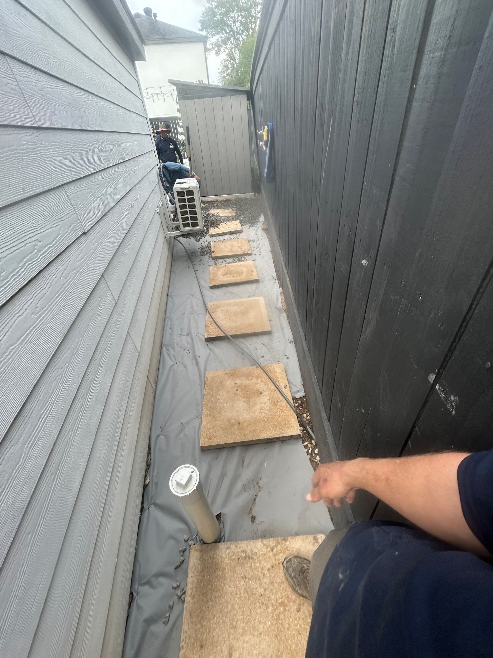 Narrow outdoor walkway with stepping stones, gray siding, black wall, and person's arm in the foreground.