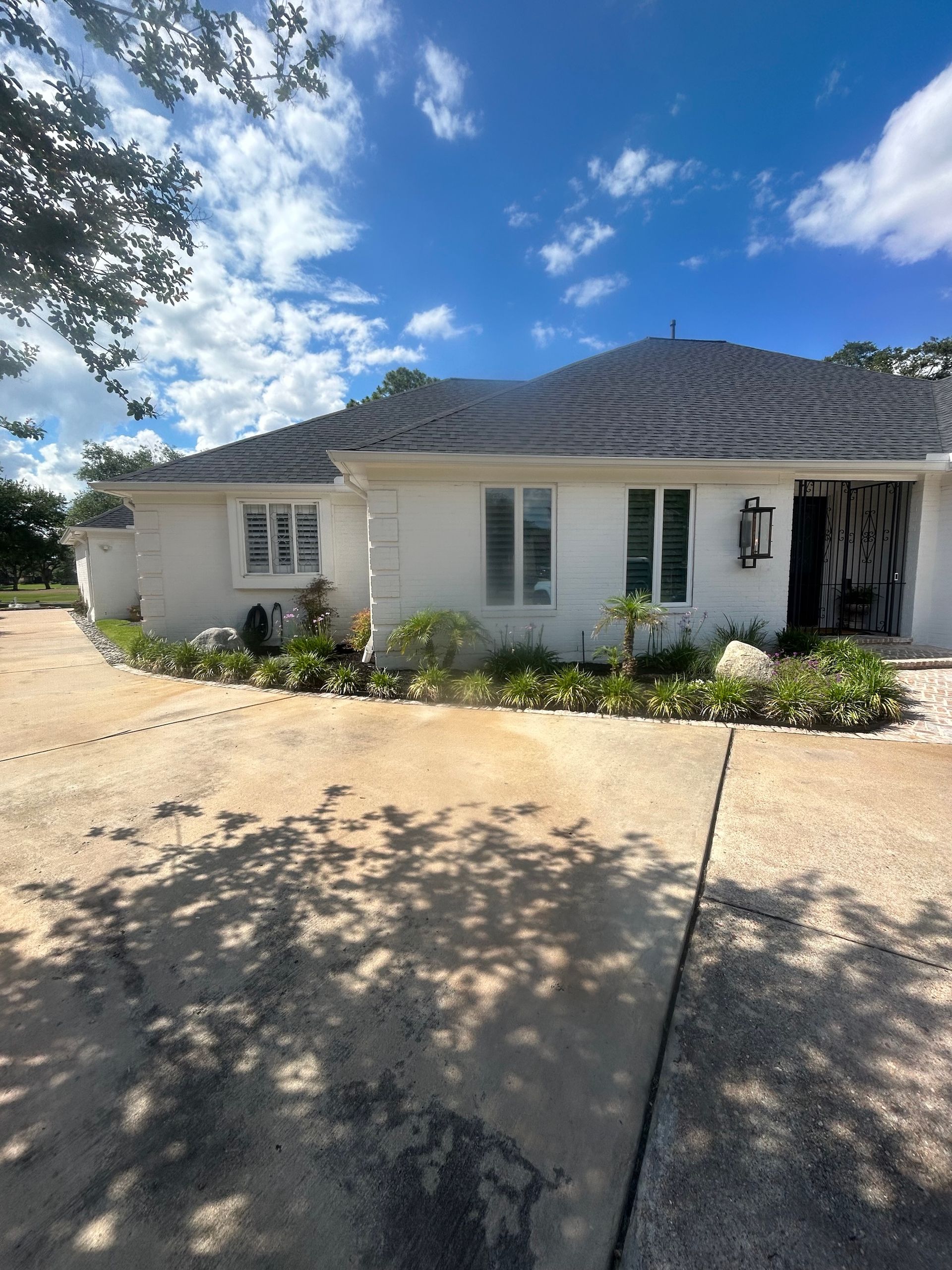 White house with a dark roof, windows, and plants along the front. A driveway leads to the house under a blue sky.