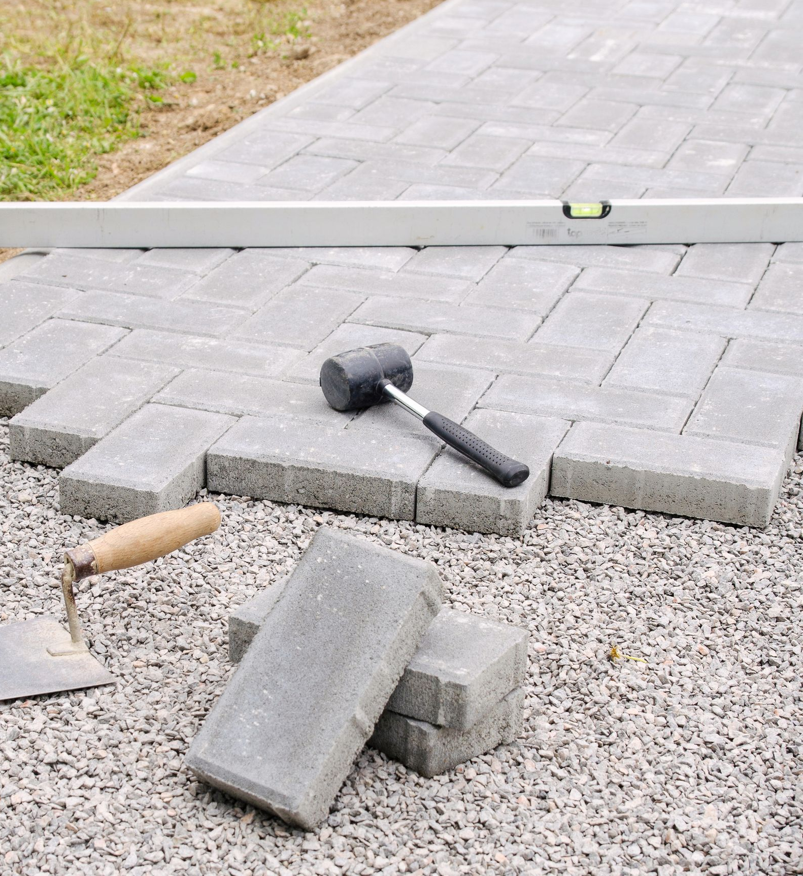 Pavers being laid on a gravel surface with a level, rubber mallet, and a trowel nearby.