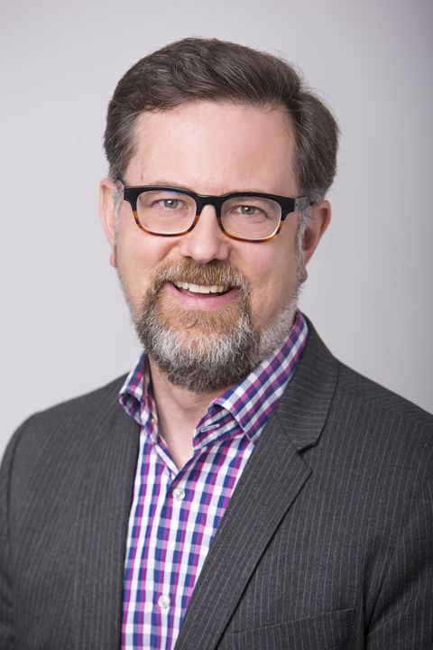 Smiling man in glasses wearing a dark blazer and purple checkered shirt against a gray background