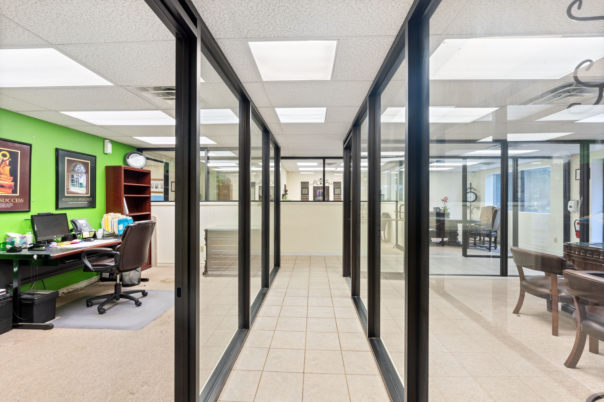 Bright office hallway with glass walls, tiled floor, and workstations visible on both sides.
