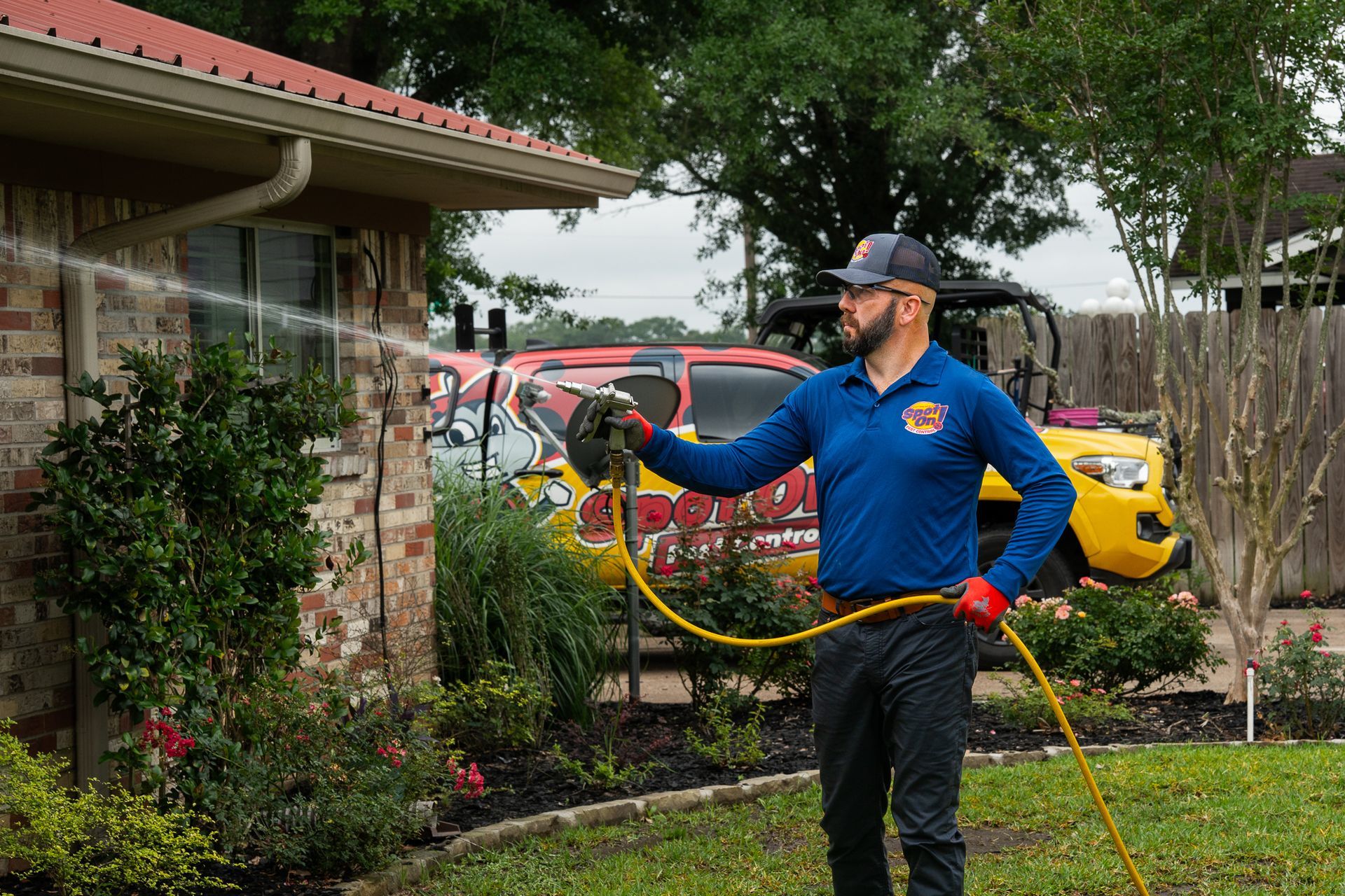 Firefighter in blue uniform spraying a hose beside a brick house, with parked cars in the background