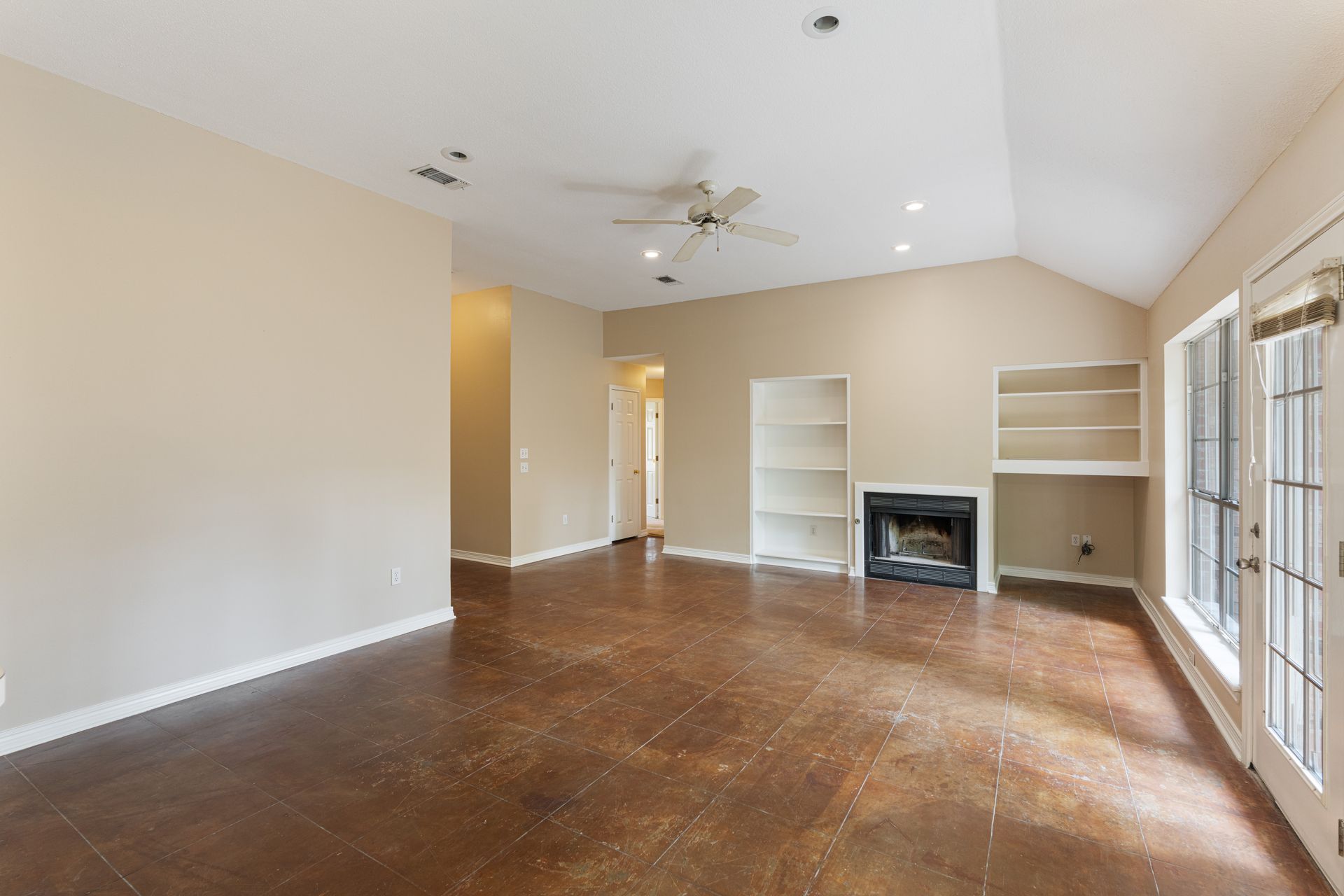Empty beige living room with hardwood floors, fireplace, built-in shelves, and French doors to the right