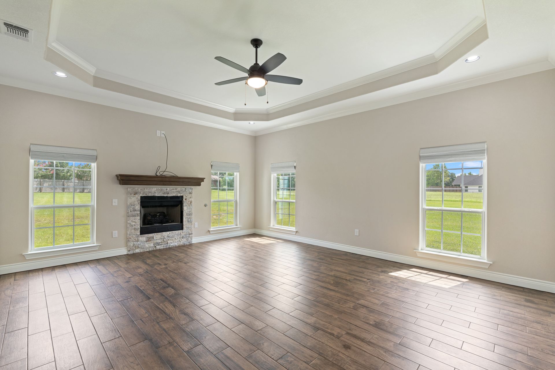 Spacious living room with dark wood floors, white walls, a fireplace, and ceiling fan overlooking a green yard