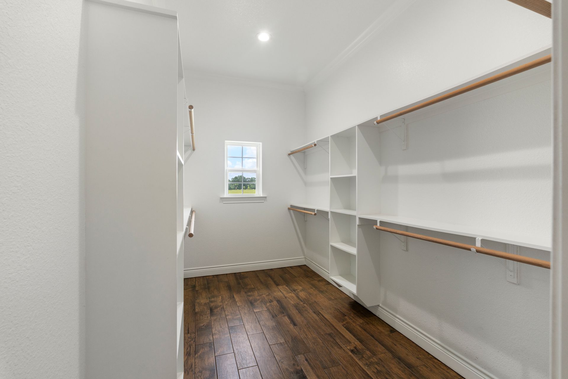 Empty walk-in closet with white shelves, hanging rods, and dark wood floors; small window at the back.